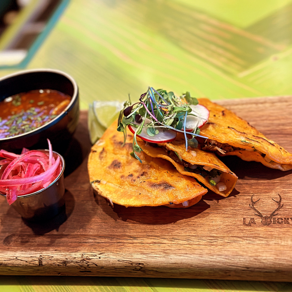 Three La Vickyna tacos with microgreens, radishes, lime, pickled onions, and a bowl of consommé.