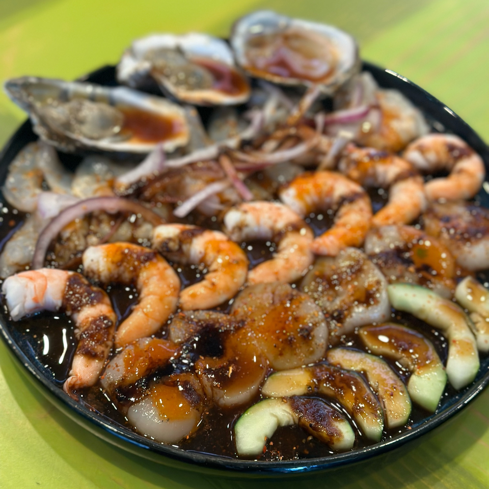 A black plate filled with shrimp, oysters, scallops, red onion, and cucumber in a dark sauce.