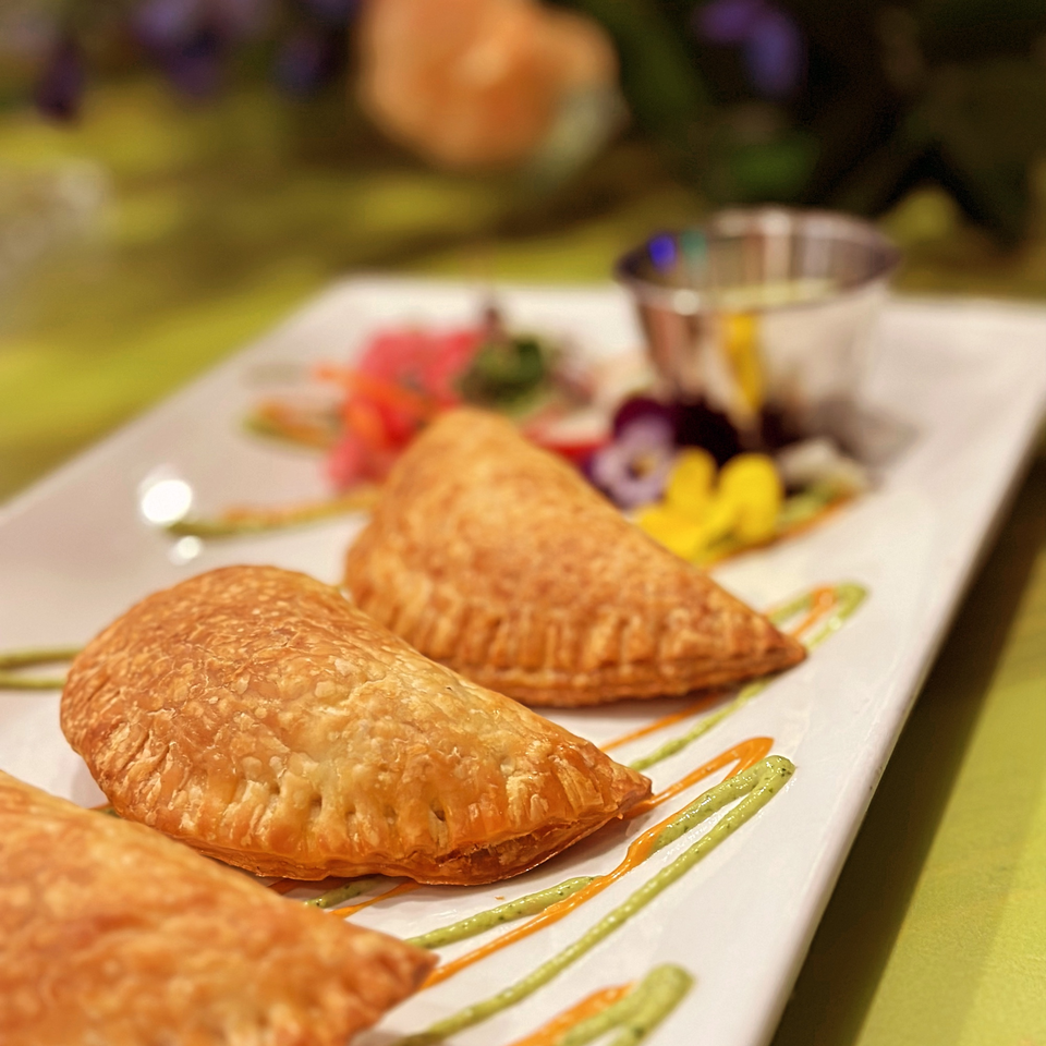 Three golden-brown empanadas on a white plate, garnished with colorful sauces and edible flowers.
